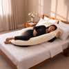 A woman in a black outfit rests on a large cream-colored U-shaped maternity pillow on a bed with white linens, in a softly lit bedroom. 49-U-Shaped Pregnancy Pillow with Customizable Head Pillow