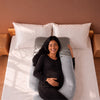 A pregnant woman lies on a bed using a light gray U-shaped maternity pillow, with white pillows and a warm brown wall in the background. 45-U-Shaped Pregnancy Pillow with Customizable Head Pillow