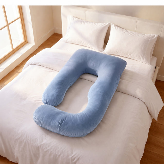 A light blue U-shaped nursing pillow resting on a neatly made bed with white linens and pillows in a bright bedroom. 46-U-Shaped Pregnancy Pillow