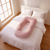 A pink U-shaped baby support pillow resting on a neatly made white bed in a bright bedroom with wooden floors and a window. 42-U-Shaped Pregnancy Pillow