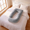 A U-shaped plush nursing pillow in a soft gray color, displayed on a neatly made bed with white bedding in a bright, sunlit bedroom. 44-U-Shaped Pregnancy Pillow