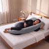 A woman lies on a bed using a large U-shaped maternity pillow in a softly lit bedroom. The pillow is light blue-gray and wraps around her body, with white bedding, a wooden bed frame, and a bedside table with flowers in the background. 43-U-Shaped Pregnancy Pillow