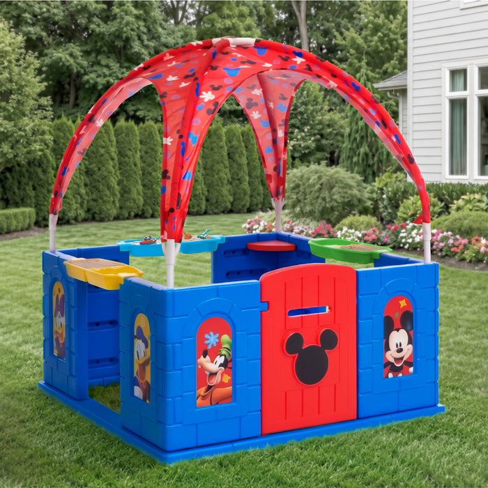 A blue and red plastic children's play yard with a curved red canopy, set up on a green lawn in front of a house. The play yard features cartoon character windows and a red door with a black mouse head silhouette. 0-Mickey Mouse Deluxe Playhouse with Canopy