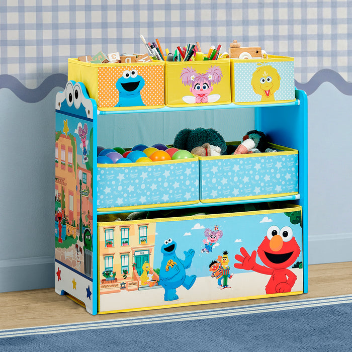 A children's storage organizer with colorful cartoon graphics, including Cookie Monster, Elmo, and Abby Cadabby, filled with toys and storage bins on a light wood floor against a blue wall. 0-Sesame Street 6 Bin Design and Store Toy Organizer
