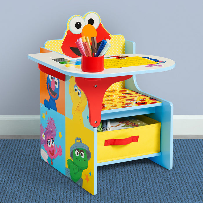 A colorful children's desk featuring Elmo on the backrest and cartoon character panels on the sides, with a red cup holding crayons on the desktop and a yellow storage drawer underneath, set against a light gray wall and blue carpet. 1-Sesame Street Chair Desk with Storage Bin