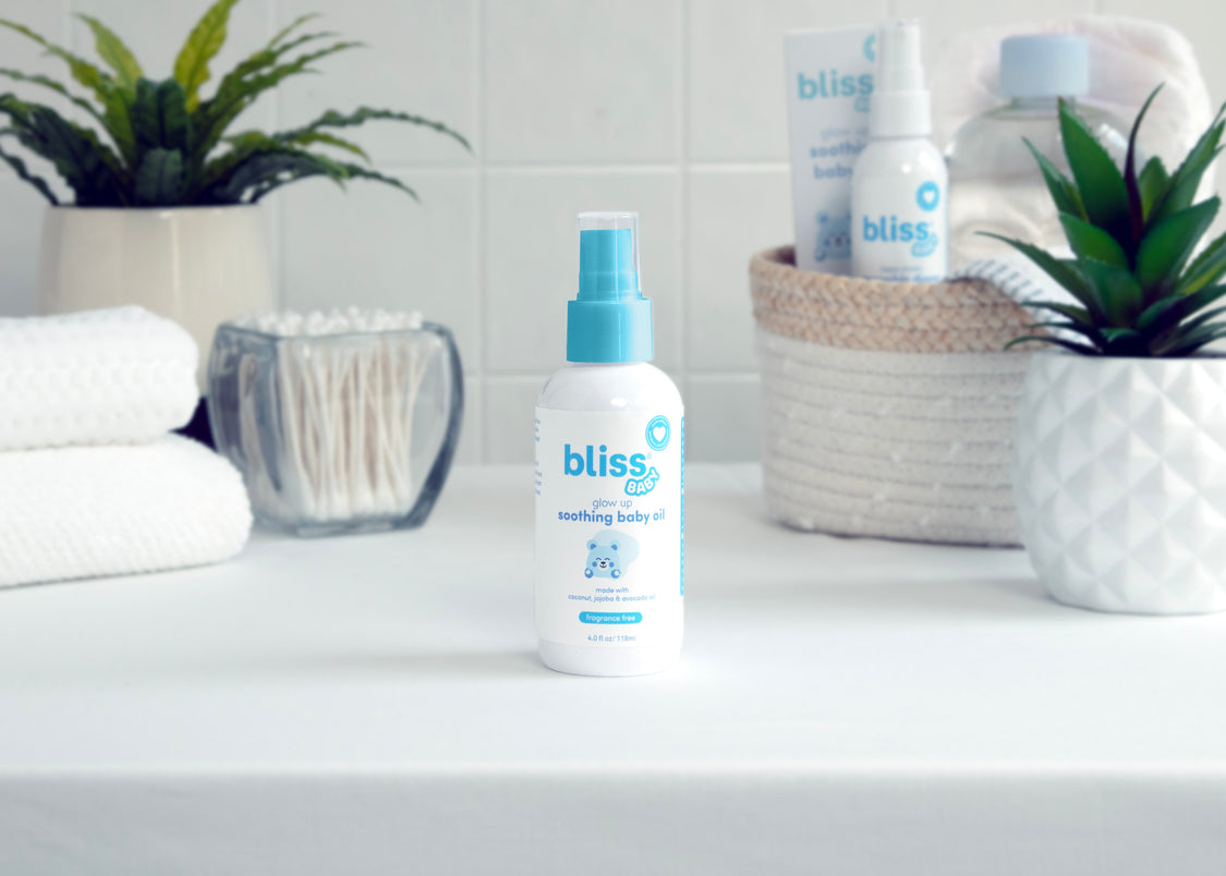 A bottle of Bliss Glow Up Soothing Baby Oil displayed on a bathroom countertop surrounded by various home décor items.
