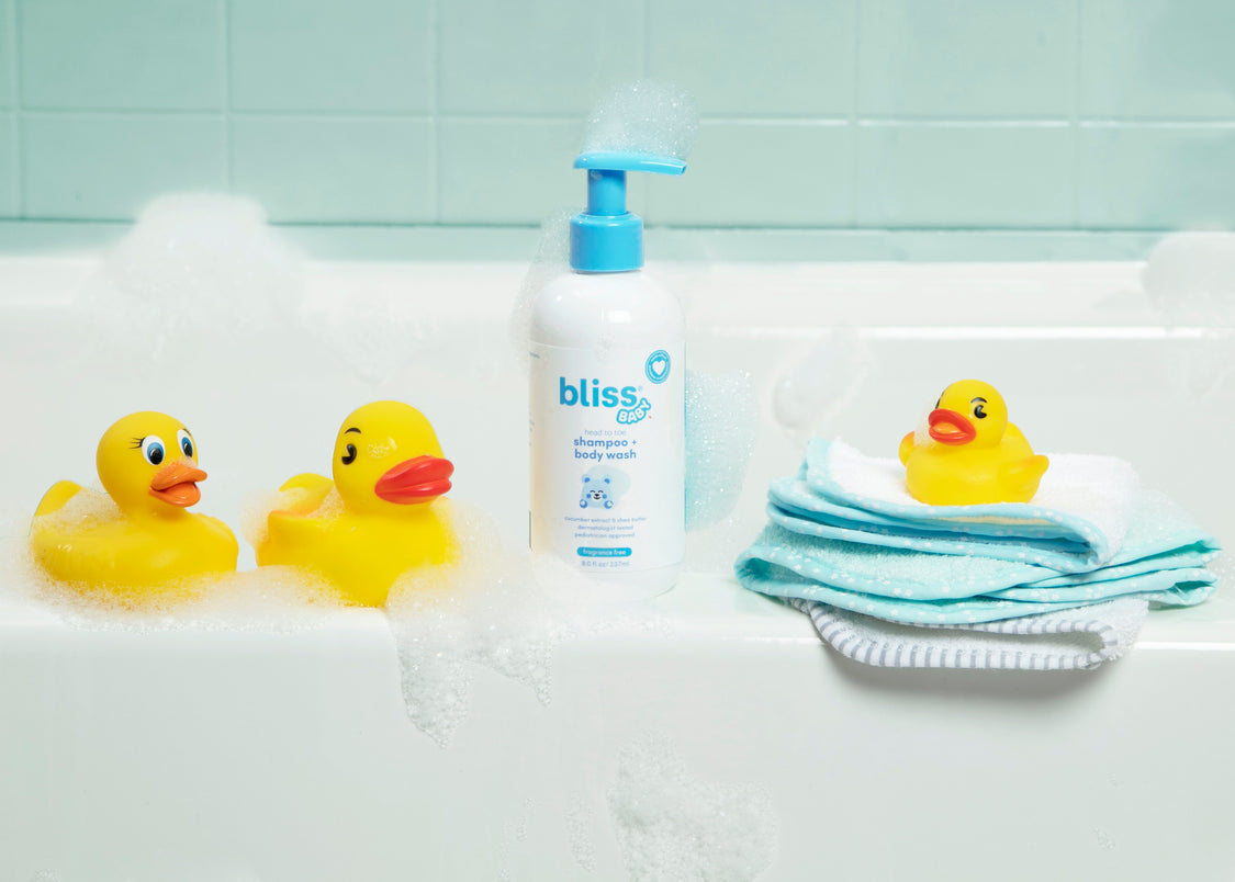 A bottle of 'Bliss' baby shampoo and body wash on a bathtub surrounded by yellow rubber ducks and a blue washcloth.