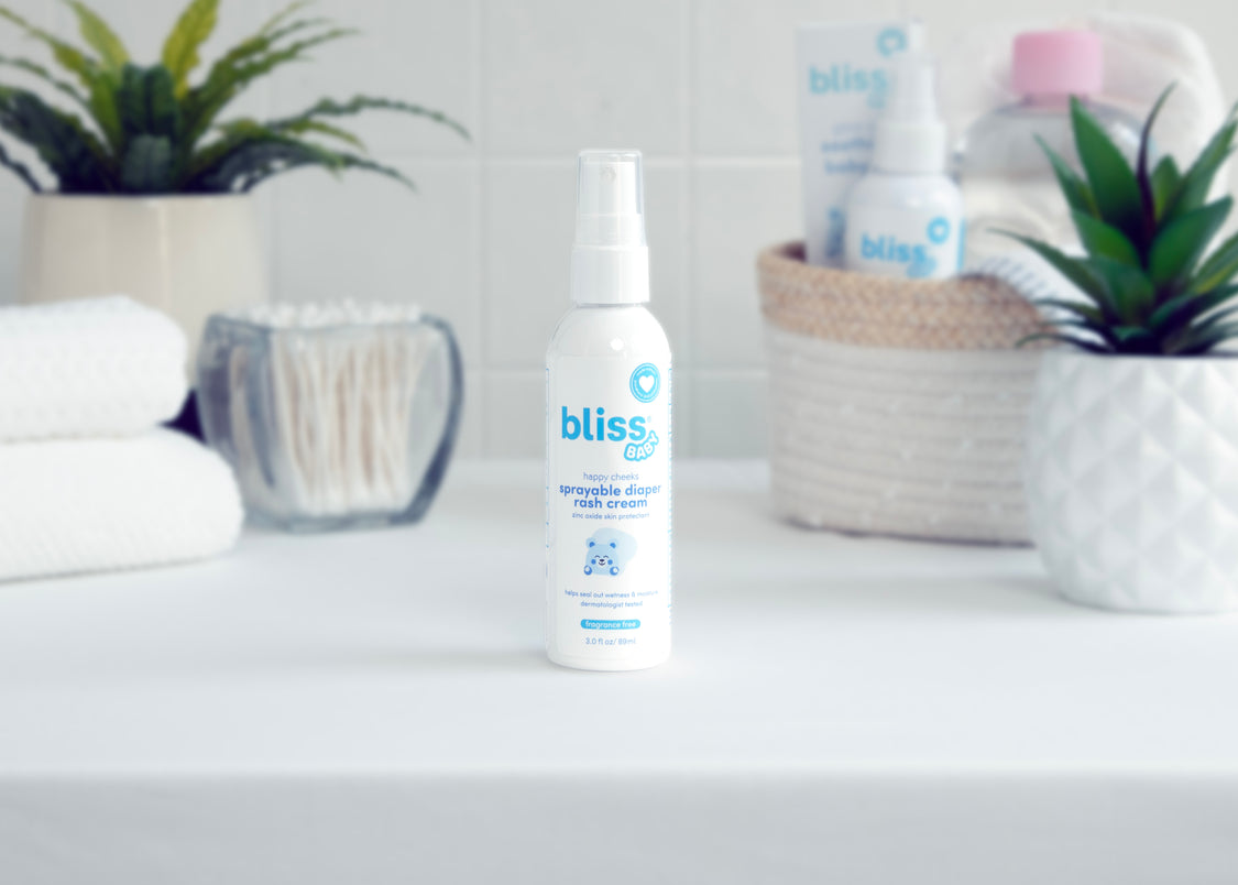 A bottle of bliss Happy Cheeks Sprayable Diaper Rash Cream placed on a bathroom shelf with blurred background elements including plants and other skincare products.