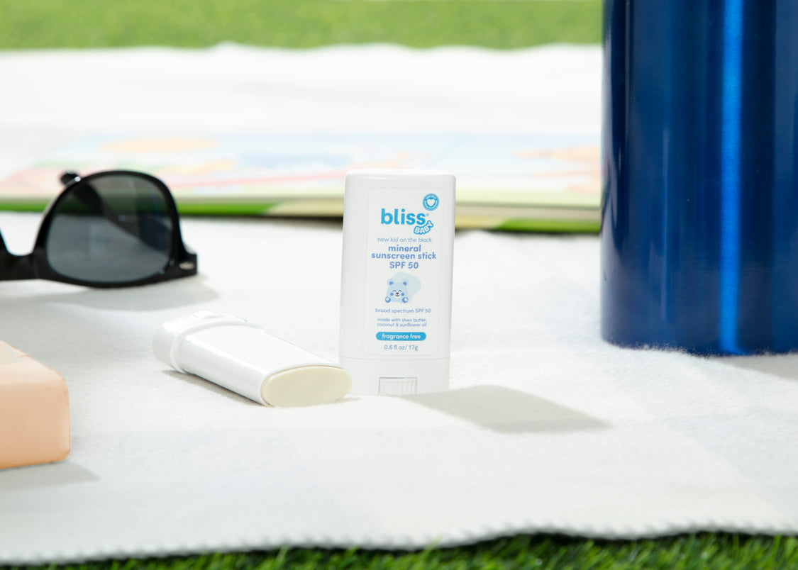 A sunscreen stick with the brand 'Bliss' placed on a grassy surface with a pair of sunglasses and a blue water bottle in the background.