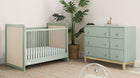 A nursery set including a crib and dresser in white and green color, with a glider and plant in the background. 1-babyGap Liam Collection