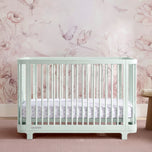 A pale mint green convertible crib with vertical slats, shown with a striped mattress and matching crib sheet, set against a pink floral and butterfly wallpaper background. - Light Sage (389) - Accessories