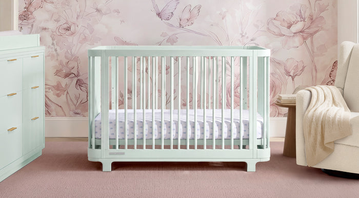 A pale mint green convertible crib with vertical slats, shown in a styled nursery setting with floral wallpaper, a matching dresser, and a cream armchair. 1-Nest Light Sage Collection