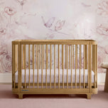 A light natural wood convertible crib with vertical slats, shown with a white and pink floral mattress. The crib is positioned in front of a pink floral wallpaper featuring butterflies and roses. - Acorn (755) - Accessories