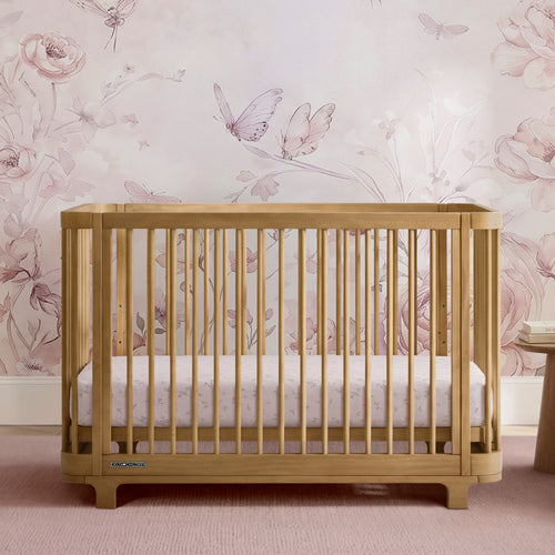 A light natural wood convertible crib with vertical slats, shown with a white and pink floral mattress. The crib is positioned in front of a pink floral wallpaper featuring butterflies and roses.