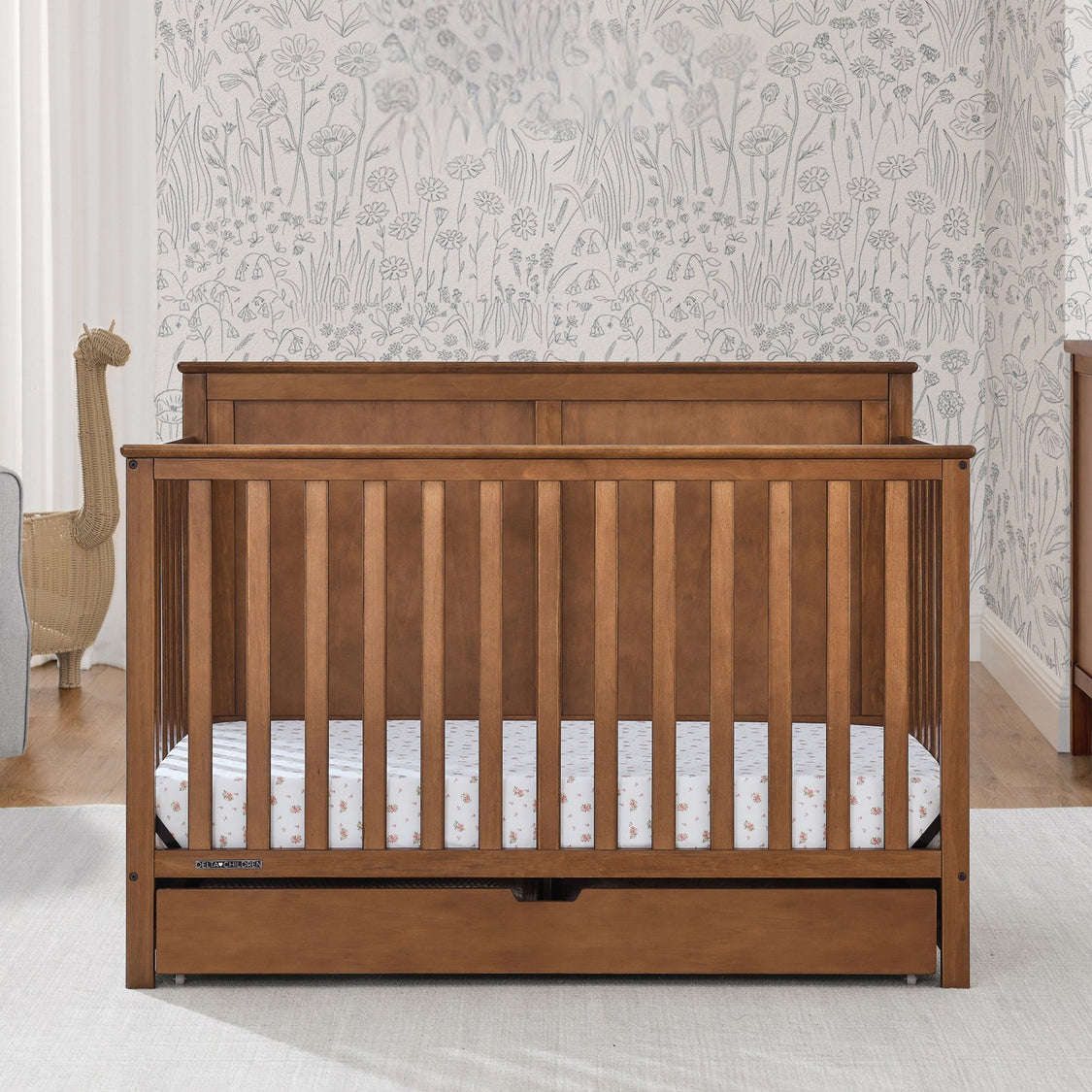 Wooden crib with a white mattress in a room with floral wallpaper.
