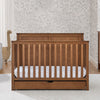 Wooden crib with a white mattress in a room with floral wallpaper. 61-Logan 6-in-1 Convertible Crib with Underdrawer