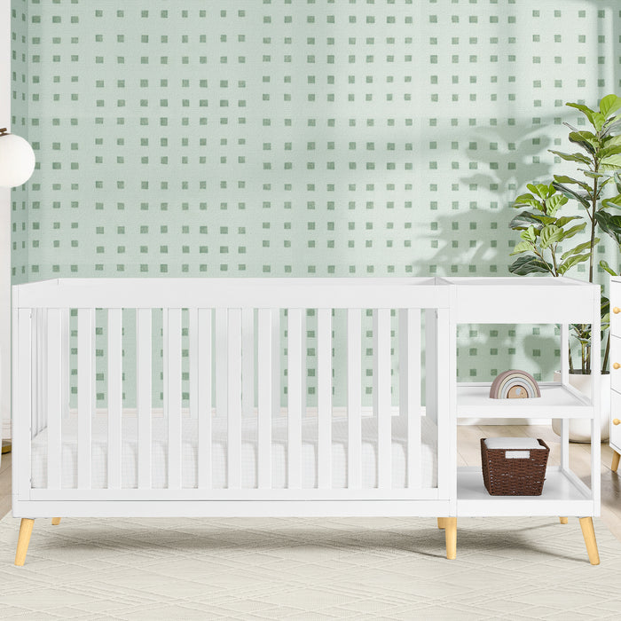 A white Essex 4-in-1 Convertible Crib and Changer with curved design and wooden accents, placed in a room with a green wall and furniture. 1-Essex 4-in-1 Convertible Crib and Changer