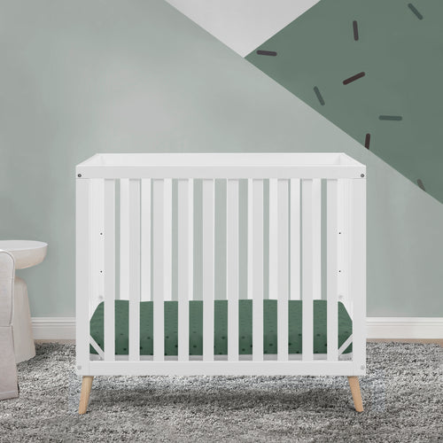 White crib with green mattress cover in a room with gray walls and carpet.