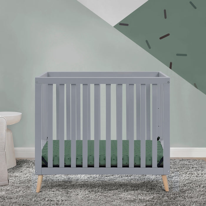 Gray crib with green mattress in a room with gray walls and carpet. 3-Essex Convertible Mini Baby Crib with 2.75-Inch Mattress, Greenguard Gold Certified