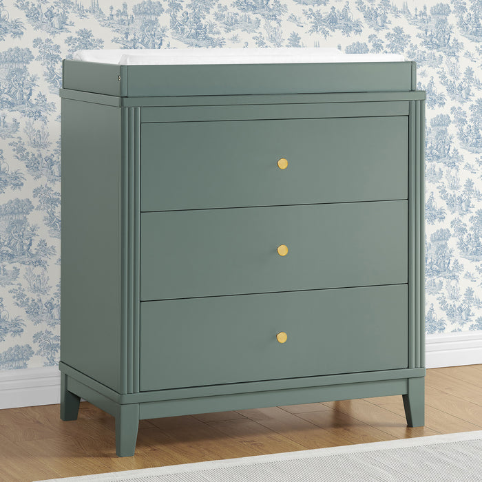 A white 3-drawer dresser with antique brass knobs and a changing top, placed against a wall with blue floral wallpaper. 1-Eloise 3 Drawer Dresser with Changing Top and Interlocking Drawers