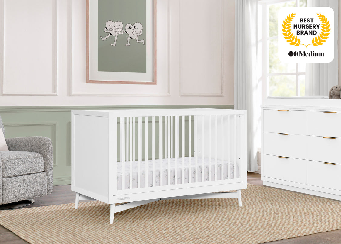 Nursery room with white crib, dresser, and gray chair, featuring a 'Best Nursery Brand' award on the wall.
