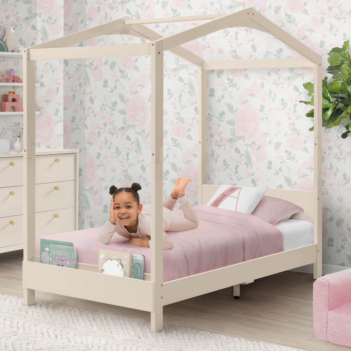 Child playing on a pink bed with a white canopy in a room with floral wallpaper.