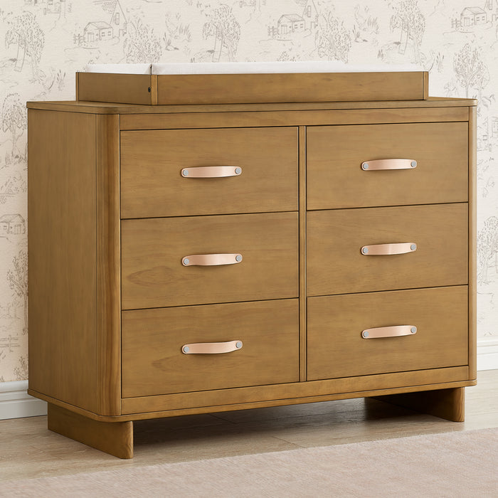 A brown wooden 6 drawer dresser with interlocking drawers, featuring leather handles and modern feet design, placed against a patterned wall. 0-babyGap Skye 6 Drawer Dresser with Interlocking Drawers