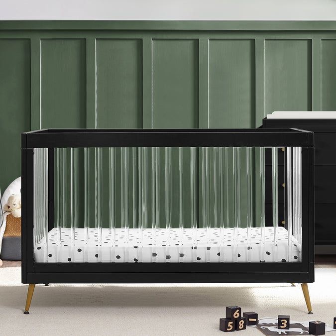 Black crib with white mattress and polka dot sheet against a green paneled wall. 17-Sloane 4-in-1 Acrylic Convertible Crib