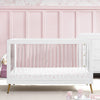 White crib with pink floral mattress cover against a pink paneled wall. 10-Sloane 4-in-1 Acrylic Convertible Crib
