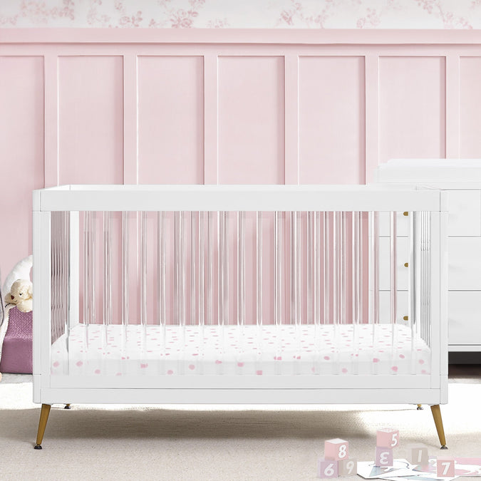 White crib with pink floral mattress cover against a pink paneled wall. 27-Sloane 4-in-1 Acrylic Convertible Crib