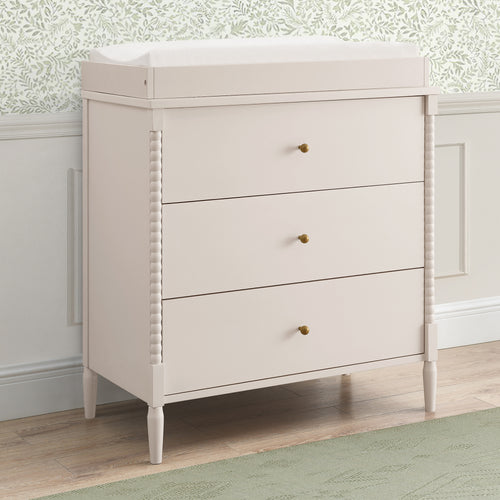 Light-colored dresser with three drawers in a room with patterned wallpaper.