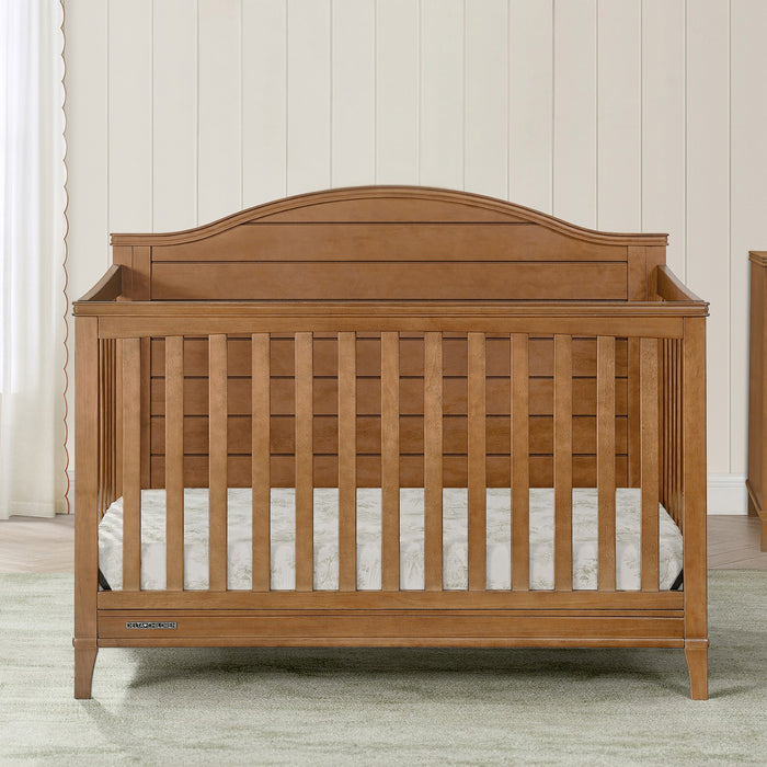 Wooden crib with slatted design in a room with light-colored walls and flooring. 3-Phoebe 6-in-1 Convertible Crib