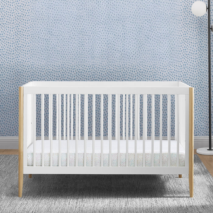 White crib with wooden sides in a room with light blue patterned wallpaper. 1-Casey 6-in-1 Convertible Crib
