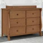 A six-drawer wooden dresser in a warm medium brown finish, featuring curved legs and round knobs, with a white changing pad mounted on top, set against a light gray wall with faint baby sketches.