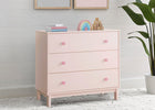 Pink dresser with three drawers in a room setting with a plant and framed pictures. 17-babyGap Legacy 3 Drawer Dresser with Interlocking Drawers