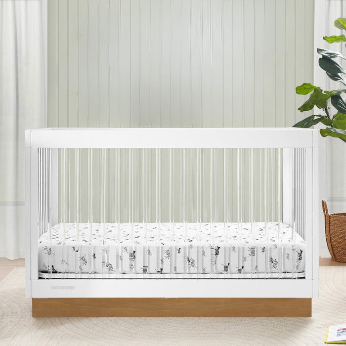 White crib with a patterned mattress in a room with light-colored walls and a plant. 2-James Acrylic 4-in-1 Convertible Crib