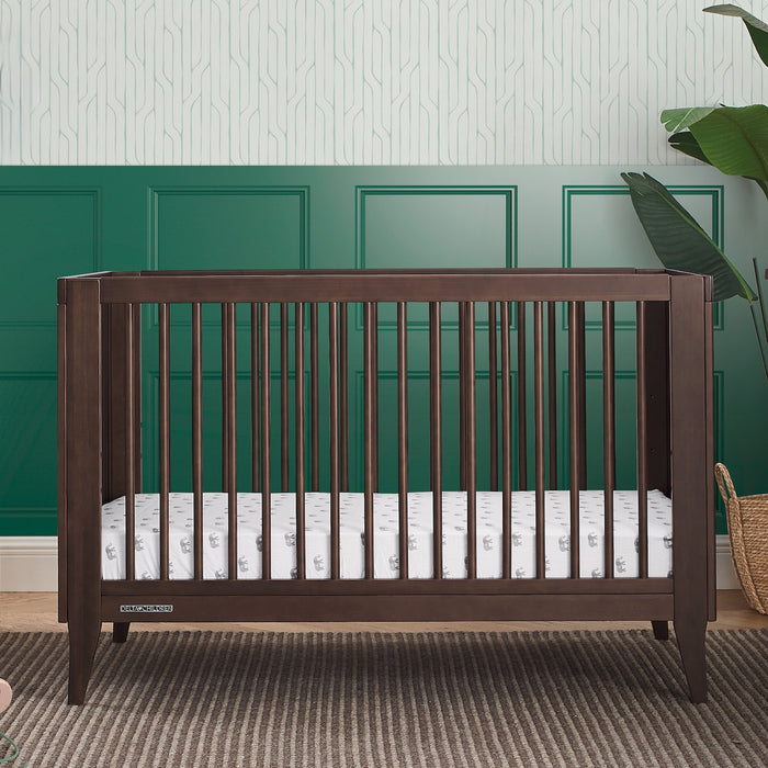 Wooden crib with white mattress against a green paneled wall 1-Ollie 4-in-1 Convertible Crib