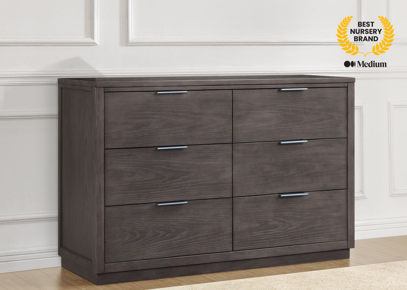 A six-drawer dresser in a dark brown wood grain finish with silver metal handles, set against a white paneled wall and light wood floor. A gold 'Best Nursery Brand' badge is visible in the top right corner.
