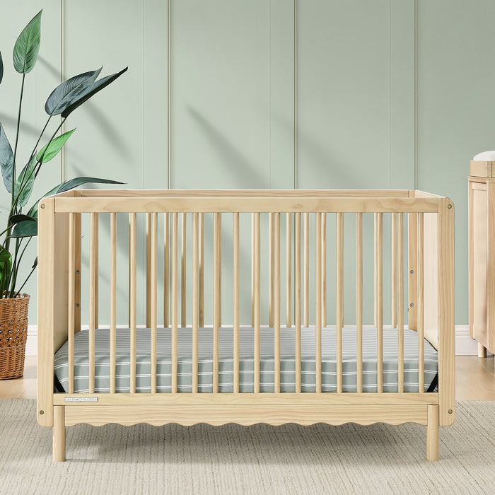 A William 4-in-1 Convertible Crib in beige color with scallop detailing, round spindles, and a light-colored dresser and chair in the background in a nursery room. 3-William 4-in-1 Convertible Crib - Greenguard Gold Certified