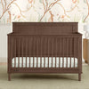 A brown Penelope 6-in-1 Convertible Crib made of sustainable pine with a satin glaze finish, shown in a nursery room with matching furniture and a decorative wall paper. 38-Penelope 6-in-1 Convertible Crib