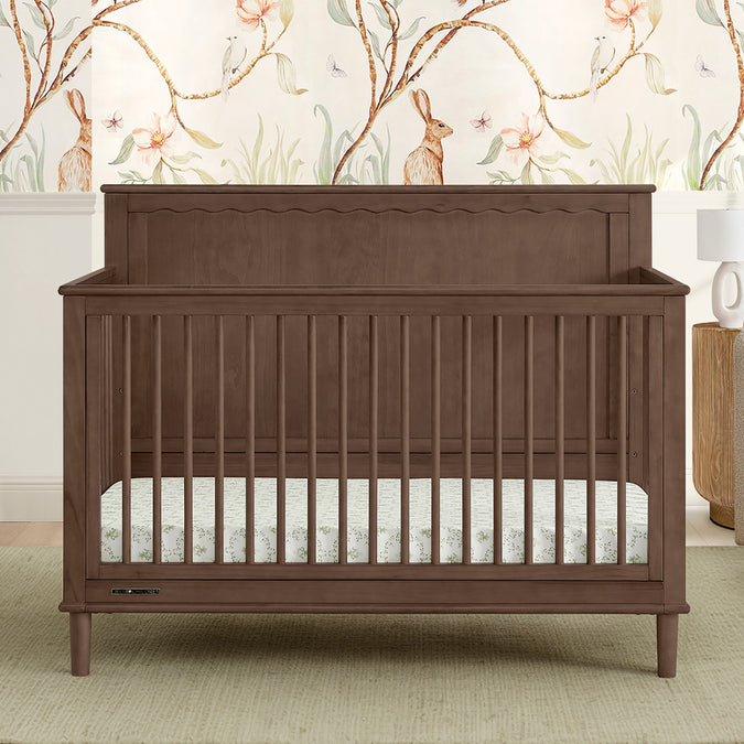 A brown Penelope 6-in-1 Convertible Crib made of sustainable pine with a satin glaze finish, shown in a nursery room with matching furniture and a decorative wall paper. 38-Penelope 6-in-1 Convertible Crib