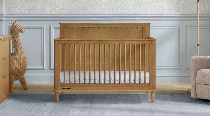 A light oak wooden convertible crib with a scalloped headboard and vertical slats, shown in a styled nursery setting with blue paneled walls, a wicker giraffe decor, and a beige armchair. 3-Penelope Collection