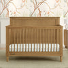 A brown Penelope 6-in-1 Convertible Crib made of sustainable pine with a satin glaze finish, shown in a nursery room with matching furniture and a decorative wall paper. 19-Penelope 6-in-1 Convertible Crib