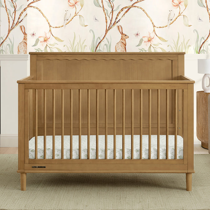 A brown Penelope 6-in-1 Convertible Crib made of sustainable pine with a satin glaze finish, shown in a nursery room with matching furniture and a decorative wall paper. 19-Penelope 6-in-1 Convertible Crib