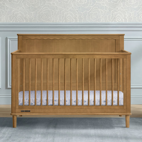 A wooden baby crib with a warm honey-oak finish, featuring a decorative scalloped headboard and vertical slats, shown in a styled nursery setting.