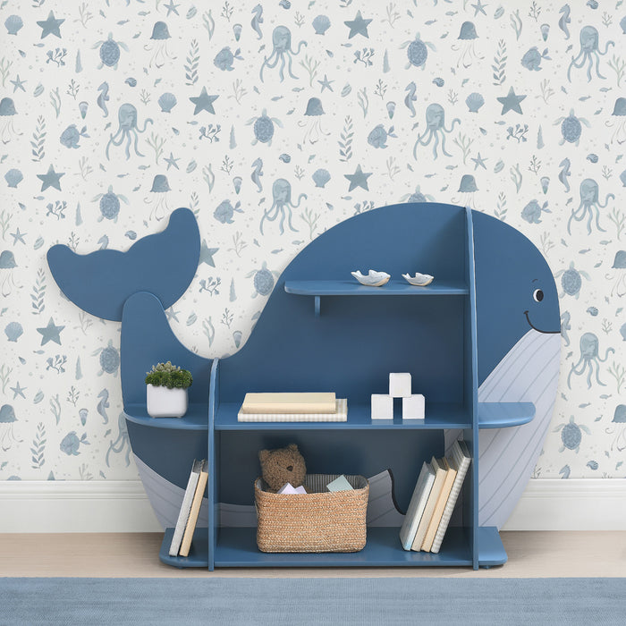 A blue whale-shaped bookcase with seven shelves, filled with books and decorative items, positioned in a room with wall paper featuring sea creatures. 1-Whale Bookcase