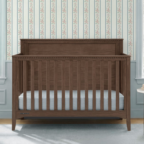 A dark brown wooden convertible crib with vertical slats and a high headboard, shown with a light blue striped mattress in a nursery setting with floral wallpaper.