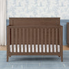 Wooden crib with white mattress in a room with floral wallpaper and gray walls. 18-Kendall 6-in-1 Convertible Crib