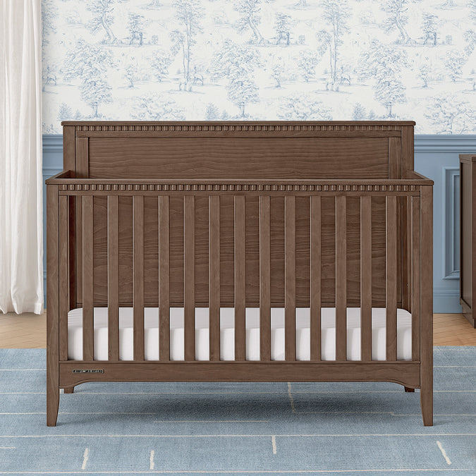 Wooden crib with white mattress in a room with floral wallpaper and gray walls. 18-Kendall 6-in-1 Convertible Crib