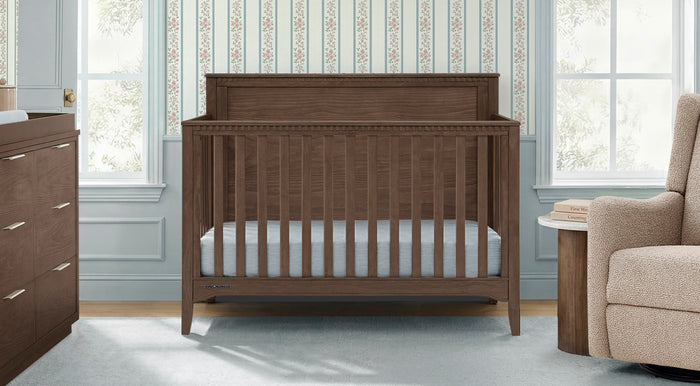 A dark brown wooden crib with a white mattress, positioned in a styled nursery setting with a matching wooden dresser on the left, a beige armchair on the right, and patterned wallpaper in the background. 2-Kendall Collection